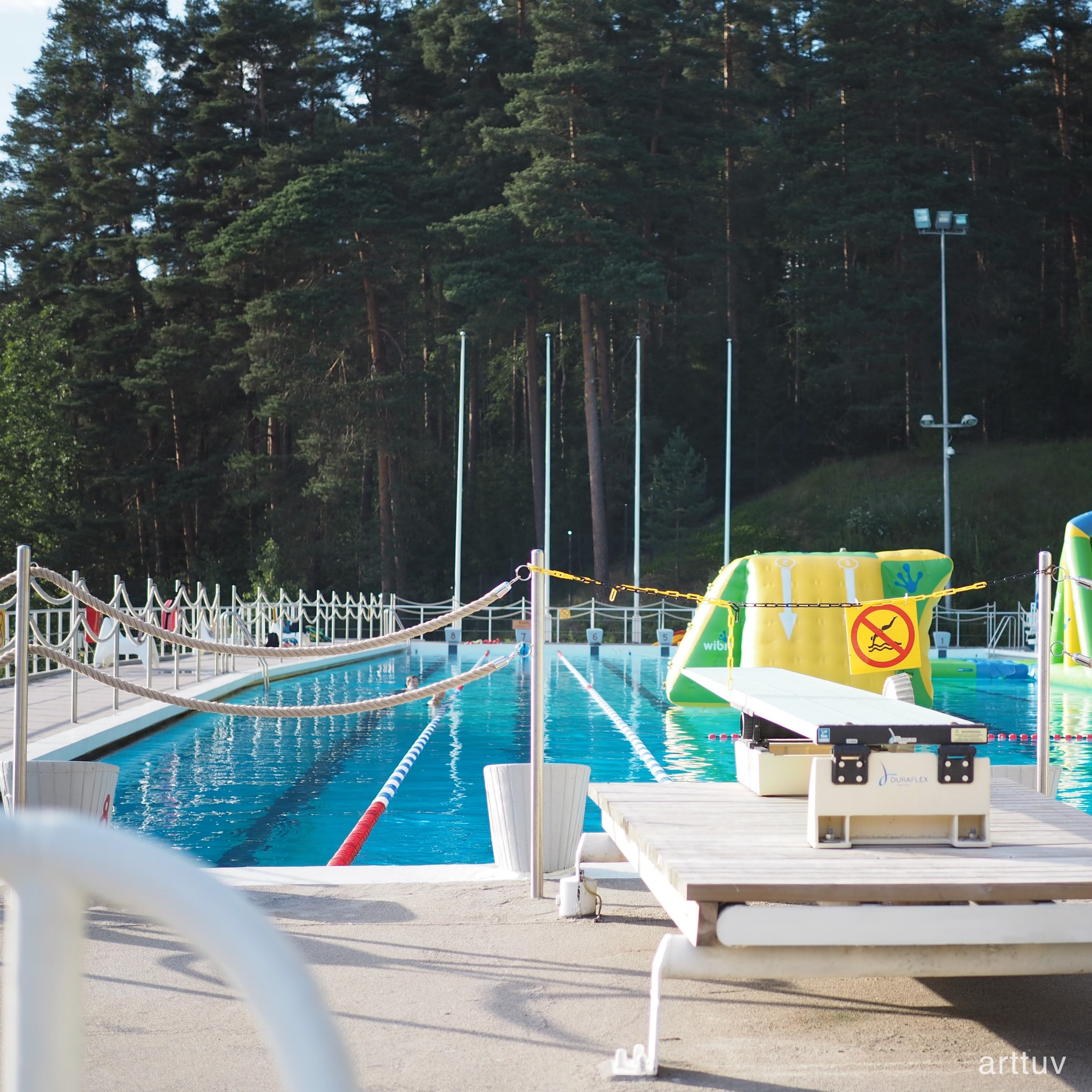 An outdoor swimming pool with lane ropes, forest in the background, and a bright inflatable play structure at the edge.