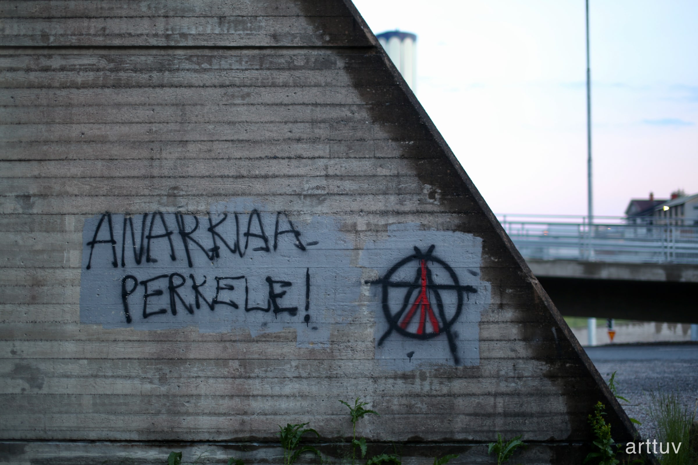 Graffiti reading Anarkia Perkele on a concrete wall, with an anarchy symbol and a bridge in the background.