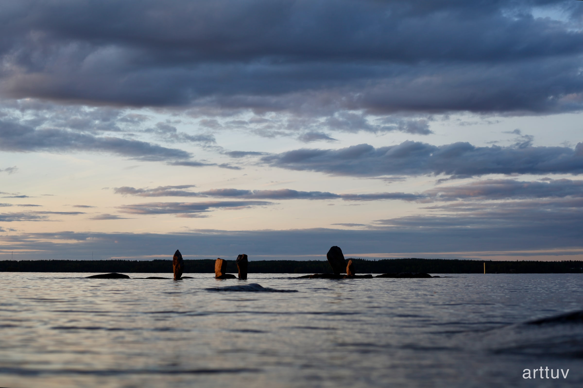 A low view across calm lake water toward upright stones beneath a wide cloudy evening sky.
