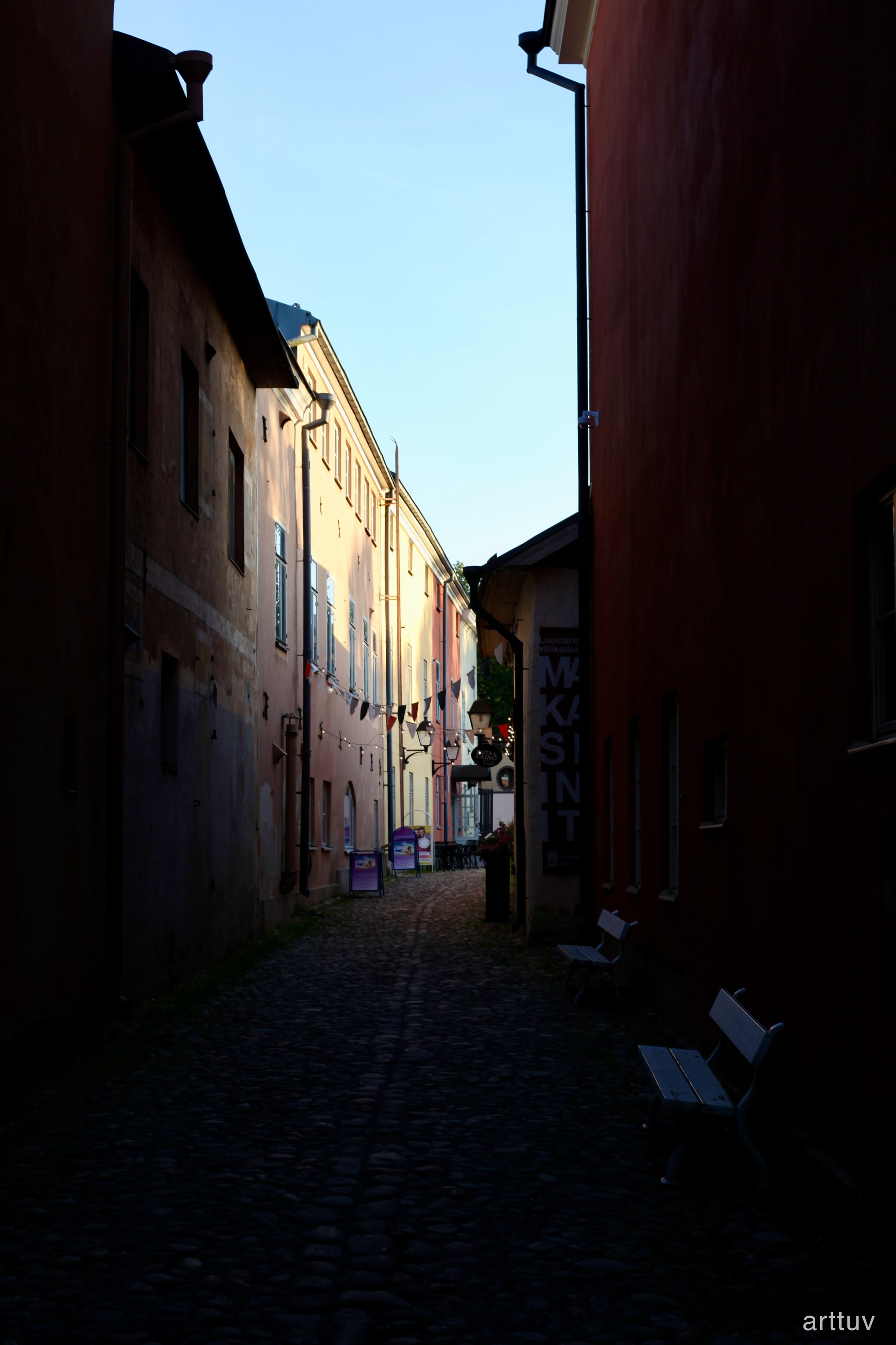 A narrow cobblestone passage between warm-toned buildings, with signs and benches in deep shade.