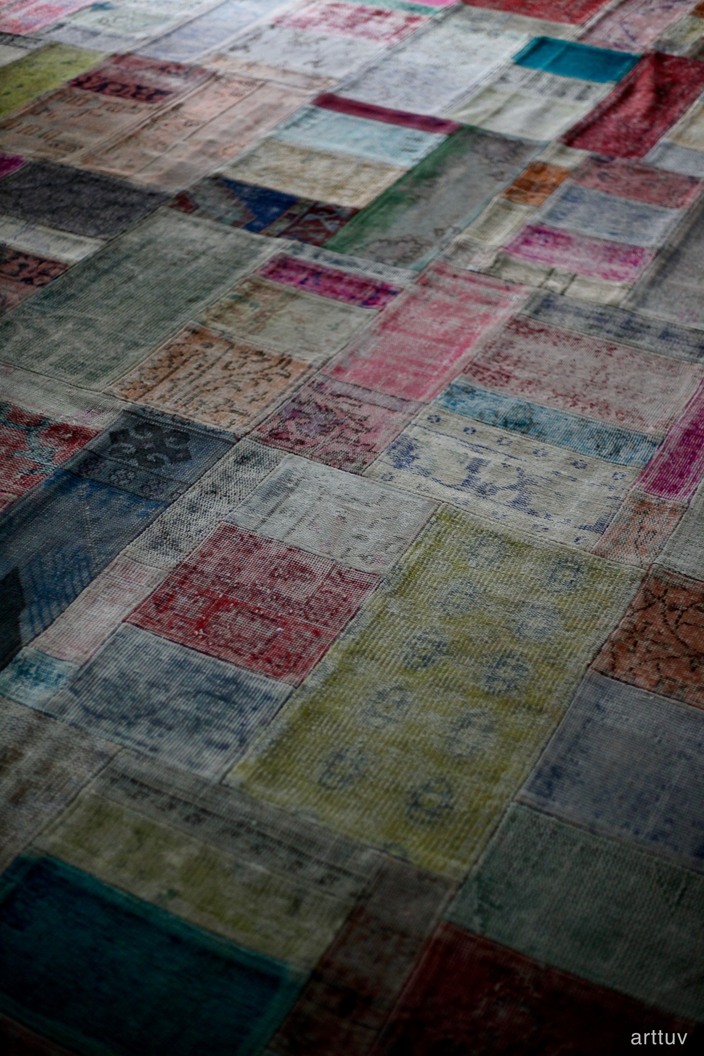 A shallow-focus photograph of a faded patchwork rug with muted red, blue, green, and cream squares.