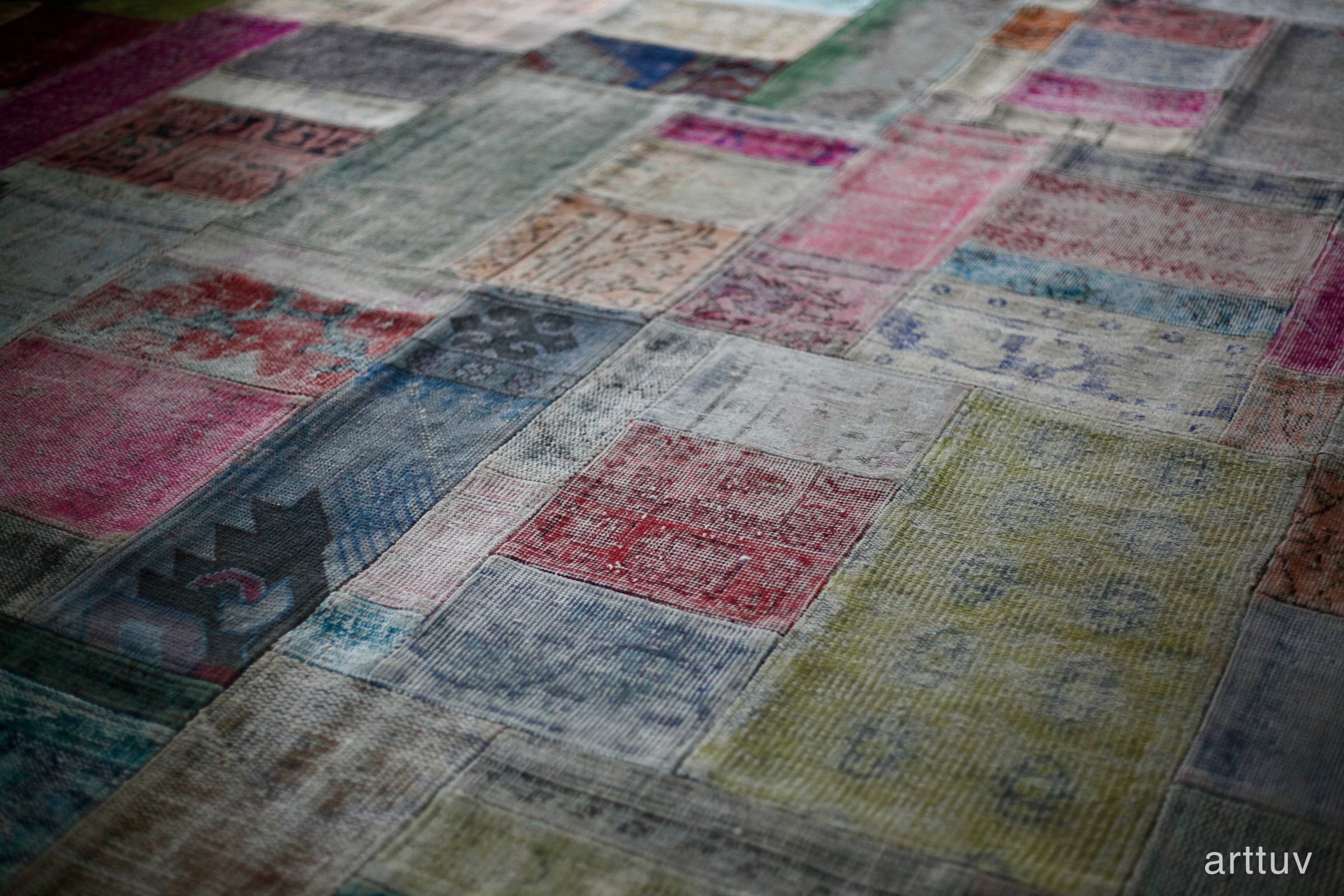 A shallow-focus photograph of a faded patchwork rug with muted red, blue, green, and cream squares.