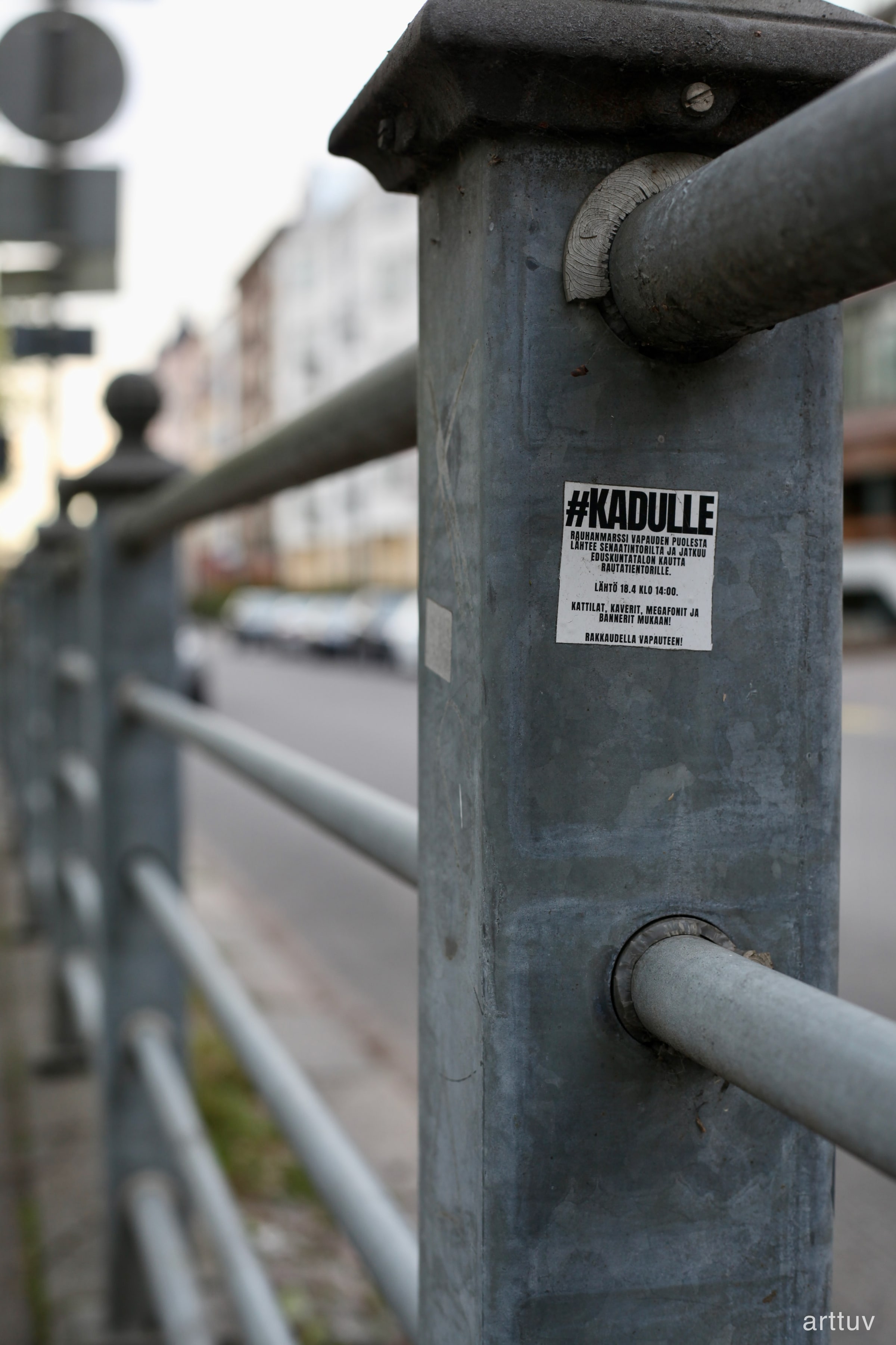 A grey metal post with a white protest sticker, photographed with a soft city background.