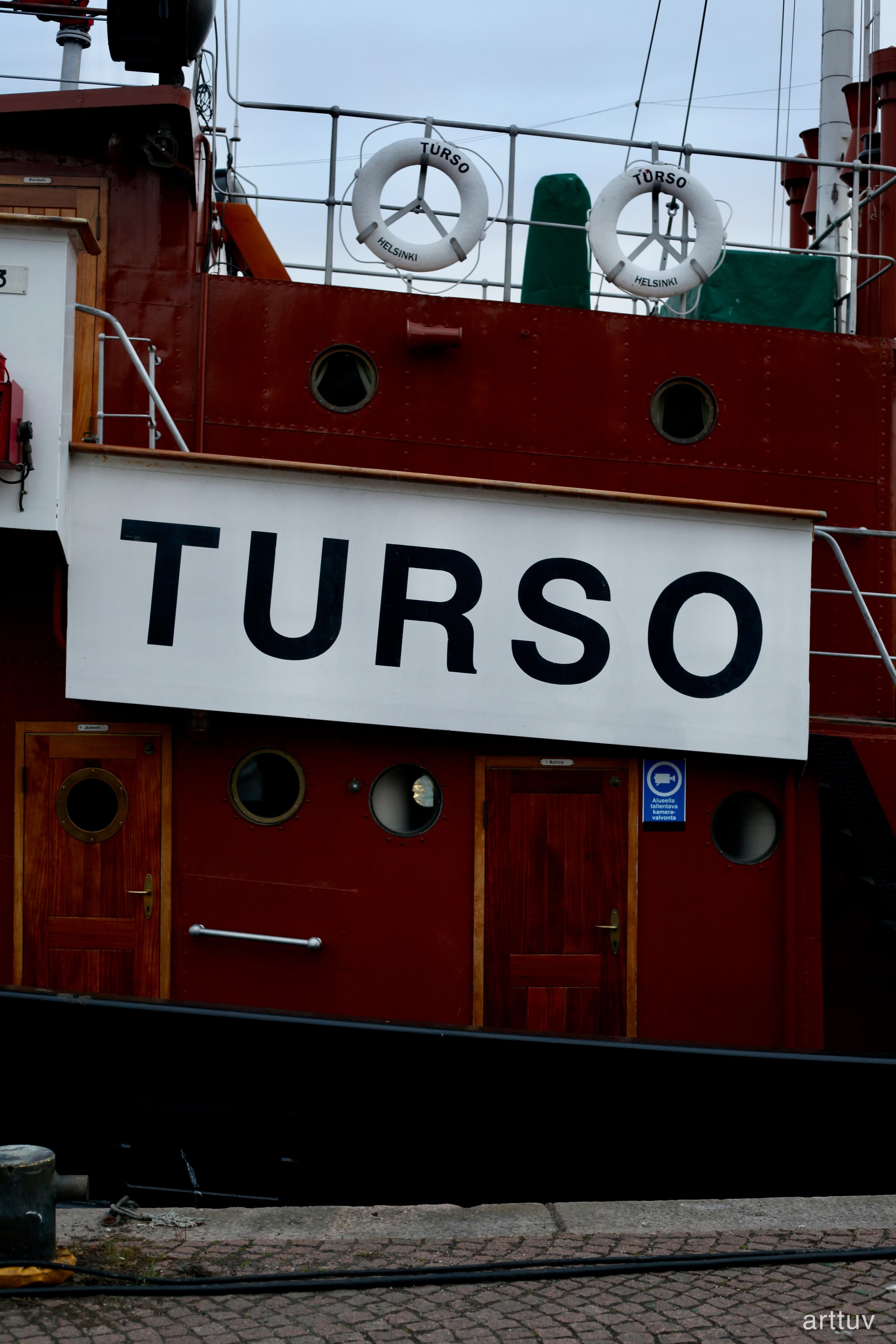 The red and white side of the ship Turso with life rings, wooden doors, and a quay in the foreground.