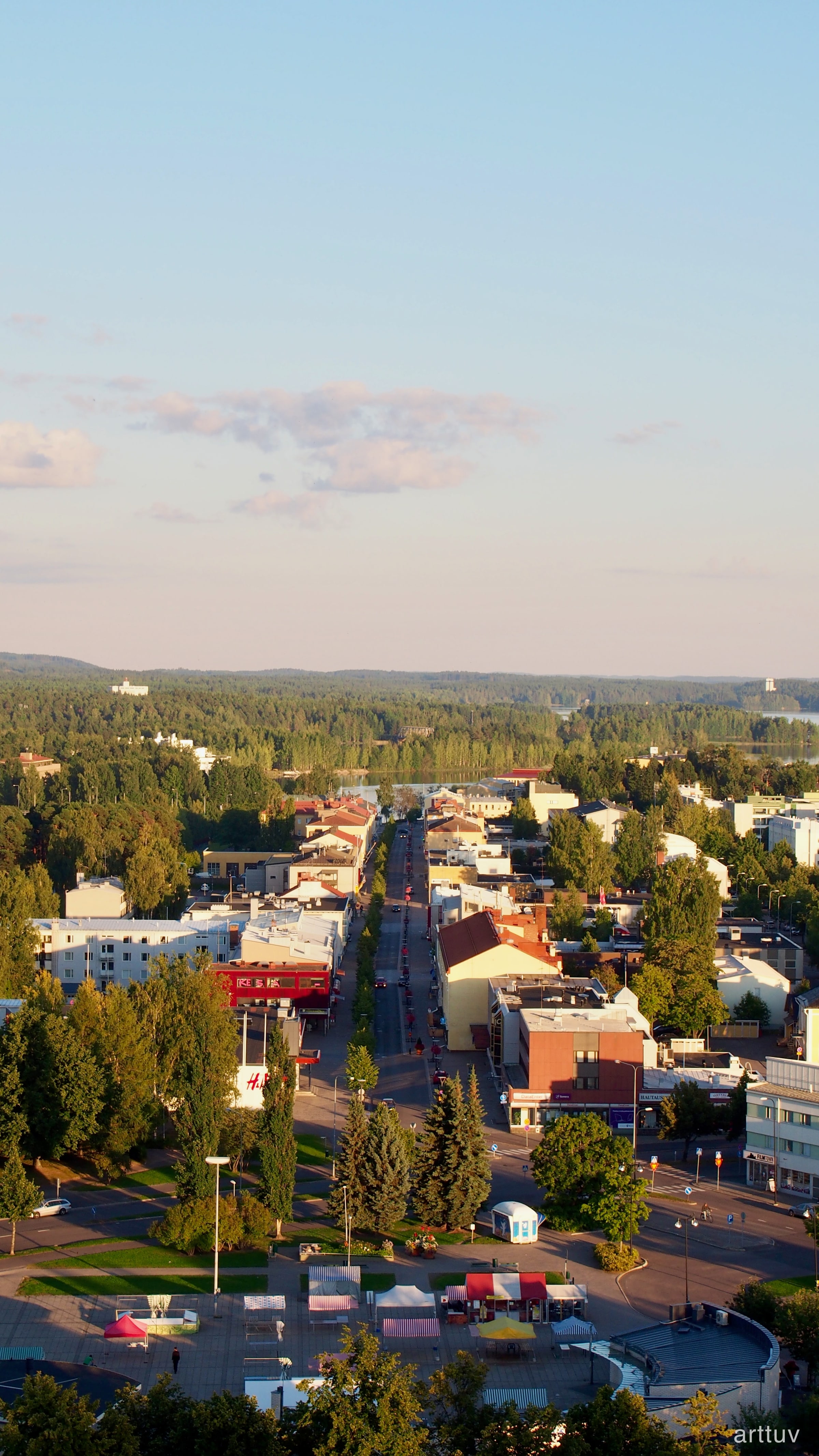 A warm evening view over a town, lake, and forest from above, with a wide pale blue sky and low clouds.